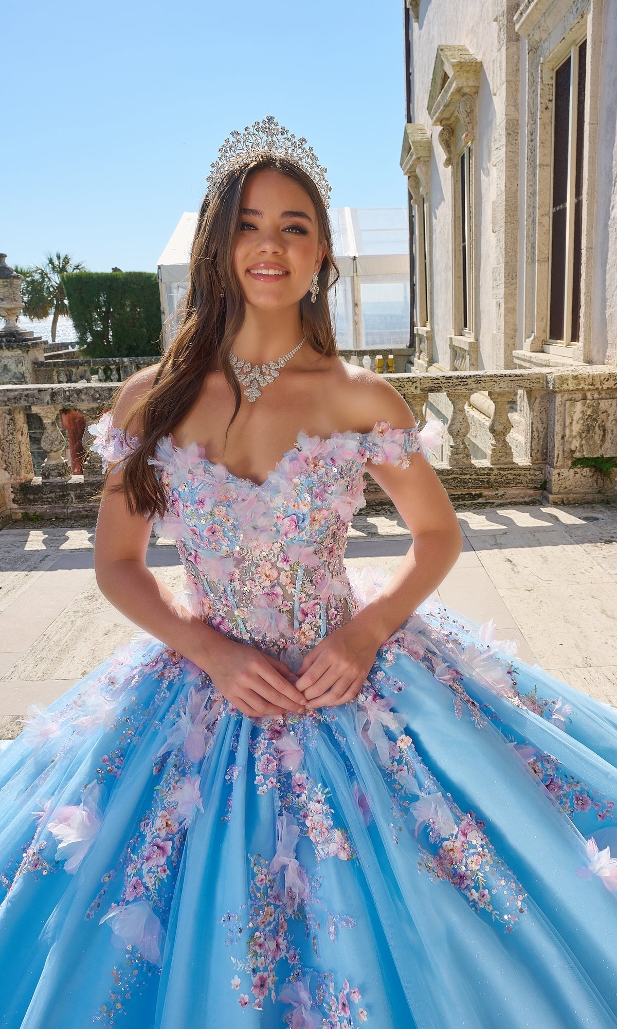 A young woman wears the Quinceanera Dress 54228 By Amarra, a blue off-the-shoulder ball gown with 3D floral appliqué, and a jeweled tiara, standing outdoors by a stone railing near a historic building.